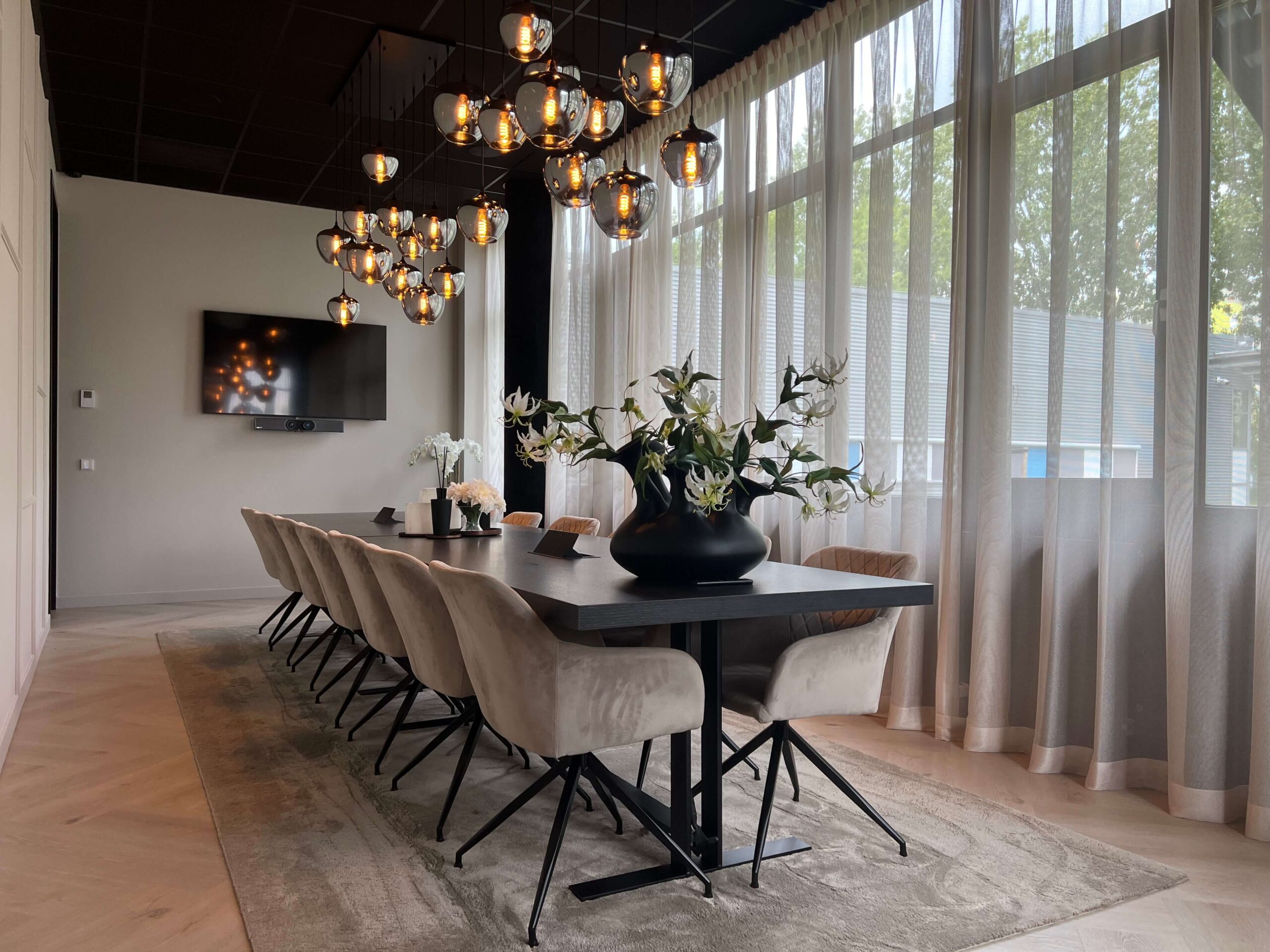 Lange eettafel met twaalf stoelen in een moderne kamer. Er hangen hanglampen boven en er staat een vaas met groene planten op tafel. Grote ramen met transparante gordijnen langs de muur links.